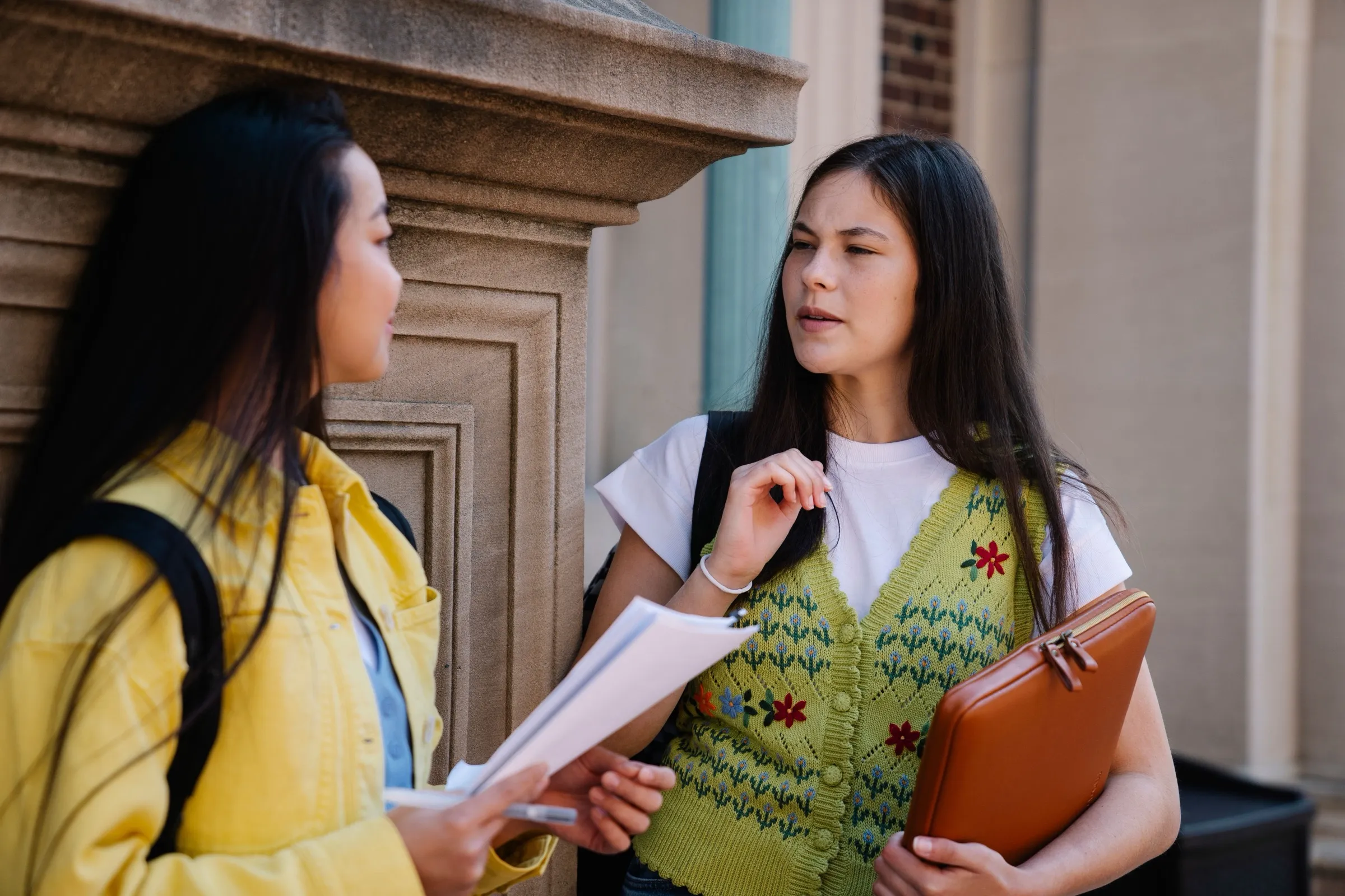 Two people in conversation beside a stone university building, holding documents